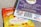 A close-up view of two packages of edible gummies on a textured surface. One package has a blue and purple design with information about 'Live Rosin Gummies,' including a 250mg THC content and '100% hemp derived' labeling. The other package is orange and yellow, labeled as 'Legal Cannabis Space Gummies' with 300mg THC content. Both packages have QR codes, branding, and legal information.