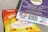 A close-up view of two packages of edible gummies on a textured surface. One package has a blue and purple design with information about 'Live Rosin Gummies,' including a 250mg THC content and '100% hemp derived' labeling. The other package is orange and yellow, labeled as 'Legal Cannabis Space Gummies' with 300mg THC content. Both packages have QR codes, branding, and legal information.