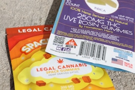 A close-up view of two packages of edible gummies on a textured surface. One package has a blue and purple design with information about 'Live Rosin Gummies,' including a 250mg THC content and '100% hemp derived' labeling. The other package is orange and yellow, labeled as 'Legal Cannabis Space Gummies' with 300mg THC content. Both packages have QR codes, branding, and legal information.