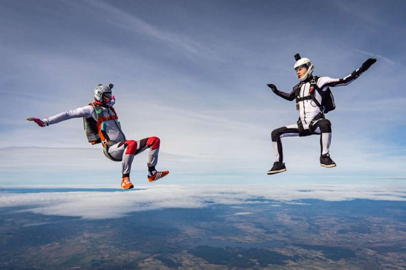 Two people in skydiving gear are captured mid-air against a backdrop of a clear sky. They are performing a synchronized freefall maneuver, with one person wearing a white suit and helmet, and the other in a gray suit with colorful accents. Beneath them, a wide expanse of land is partially visible.