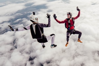 Two skydivers are captured mid-air against a backdrop of fluffy clouds. They appear to be in freefall, wearing helmets and parachute gear. One is dressed in red and the other in a black and white suit. Both individuals are positioned in a dynamic pose, giving a sense of motion and excitement.