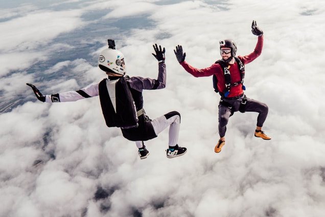 Two skydivers are captured mid-air against a backdrop of fluffy clouds. They appear to be in freefall, wearing helmets and parachute gear. One is dressed in red and the other in a black and white suit. Both individuals are positioned in a dynamic pose, giving a sense of motion and excitement.