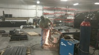 A person is using a grinding tool, creating bright sparks in an industrial workshop. The space contains various metal pipes, pallets, a blue welding machine, and shelves stocked with containers.