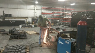 A person is using a grinding tool, creating bright sparks in an industrial workshop. The space contains various metal pipes, pallets, a blue welding machine, and shelves stocked with containers.