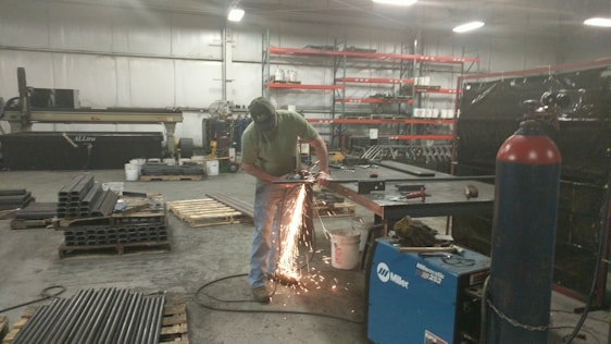 A person is using a grinding tool, creating bright sparks in an industrial workshop. The space contains various metal pipes, pallets, a blue welding machine, and shelves stocked with containers.