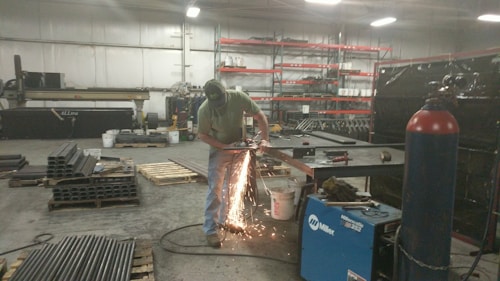 A person is using a grinding tool, creating bright sparks in an industrial workshop. The space contains various metal pipes, pallets, a blue welding machine, and shelves stocked with containers.