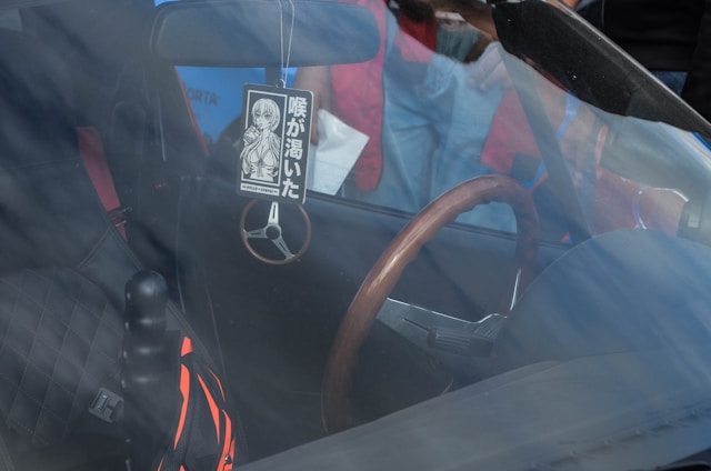 The interior of a car is visible through the windshield, featuring a wooden steering wheel and a diamond-patterned seat cover. A decorative air freshener with Japanese characters and an illustration is hanging from the rearview mirror. A striped bag is resting on the passenger seat. The reflection of people and a nearby outdoor environment can also be seen through the glass.