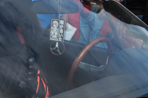 The interior of a car is visible through the windshield, featuring a wooden steering wheel and a diamond-patterned seat cover. A decorative air freshener with Japanese characters and an illustration is hanging from the rearview mirror. A striped bag is resting on the passenger seat. The reflection of people and a nearby outdoor environment can also be seen through the glass.