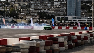 A green car is drifting around a track, creating smoke as it navigates a course bordered by red and white barriers. In the background, there are spectators gathered behind a fence, with a cityscape of tall buildings. The scene is set during daylight, with banners and flags indicating a possible event or competition.
