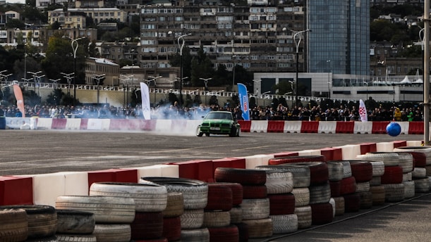 A green car is drifting around a track, creating smoke as it navigates a course bordered by red and white barriers. In the background, there are spectators gathered behind a fence, with a cityscape of tall buildings. The scene is set during daylight, with banners and flags indicating a possible event or competition.