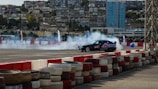 A black racing car drifts on an asphalt track surrounded by smoke. The track is enclosed with stacks of colored tires forming a barrier, and a large crowd watches in the background. Residential buildings and a tall glass skyscraper tower over the scene.