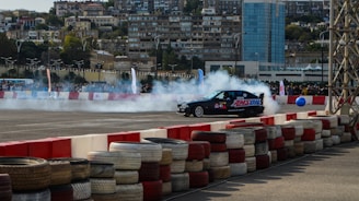 A black racing car drifts on an asphalt track surrounded by smoke. The track is enclosed with stacks of colored tires forming a barrier, and a large crowd watches in the background. Residential buildings and a tall glass skyscraper tower over the scene.