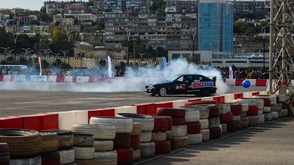 A black racing car drifts on an asphalt track surrounded by smoke. The track is enclosed with stacks of colored tires forming a barrier, and a large crowd watches in the background. Residential buildings and a tall glass skyscraper tower over the scene.
