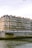 A large, classic European apartment building with multiple windows and balconies lines the water's edge. The structure exudes historical architectural charm and features a slightly worn facade. The water is calm, and a stone embankment runs parallel to the river. The sky is overcast, adding an atmospheric touch.