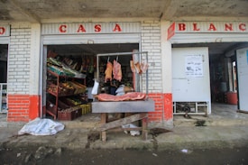 A rustic storefront displays various fresh produce in red and yellow crates to the left, with cuts of fresh meat hanging on a metal frame and lying on a wooden table in front. The shop has weathered brick walls painted with red and white, and the open design allows a glimpse into a modest, busy interior. Bags and signs are scattered around the entrance.