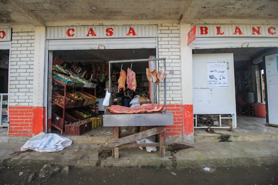 A rustic storefront displays various fresh produce in red and yellow crates to the left, with cuts of fresh meat hanging on a metal frame and lying on a wooden table in front. The shop has weathered brick walls painted with red and white, and the open design allows a glimpse into a modest, busy interior. Bags and signs are scattered around the entrance.