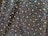 A textured fabric with an intricate pattern of small, shiny beads and larger iridescent sequins. The sequins reflect a spectrum of colors, creating a vibrant, shimmering effect against the dark background.