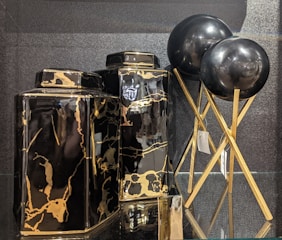 Two black and gold marble-patterned vases are displayed alongside two black spherical objects on golden stands. The vases have a glossy finish, with the marble pattern adding a luxurious touch. The spherical objects appear to be decorative, supported by elegant crisscrossed stands.