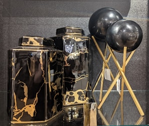 Two black and gold marble-patterned vases are displayed alongside two black spherical objects on golden stands. The vases have a glossy finish, with the marble pattern adding a luxurious touch. The spherical objects appear to be decorative, supported by elegant crisscrossed stands.