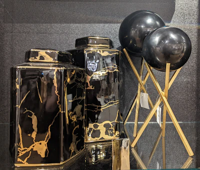 Two black and gold marble-patterned vases are displayed alongside two black spherical objects on golden stands. The vases have a glossy finish, with the marble pattern adding a luxurious touch. The spherical objects appear to be decorative, supported by elegant crisscrossed stands.