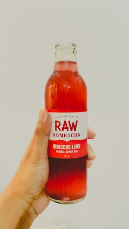 A hand holds a glass bottle labeled 'RAW Kombucha' with hibiscus lime flavoring and the text 'herbal super tea'. The bottle contains a red liquid, and the background is plain and light-colored.