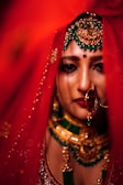 A person is adorned in ornate jewelry, featuring vibrant green and gold accents. They wear a red veil partially covering their face, with a decorative headpiece and a large nose ring. The image captures the rich texture and detail of the attire in a close-up shot.