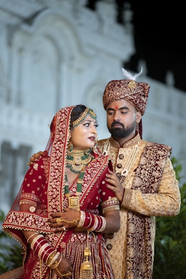 A couple dressed in traditional Indian wedding attire stands in an ornate setting. The woman is wearing a richly decorated red lehenga embellished with gold embroidery, along with intricate jewelry including a necklace, earrings, and bangles. The man is wearing a coordinated sherwani, featuring detailed embroidery and a turban. They appear poised and formal.