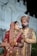A couple dressed in traditional Indian wedding attire stands in an ornate setting. The woman is wearing a richly decorated red lehenga embellished with gold embroidery, along with intricate jewelry including a necklace, earrings, and bangles. The man is wearing a coordinated sherwani, featuring detailed embroidery and a turban. They appear poised and formal.