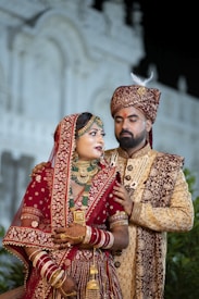 A couple dressed in traditional Indian wedding attire stands in an ornate setting. The woman is wearing a richly decorated red lehenga embellished with gold embroidery, along with intricate jewelry including a necklace, earrings, and bangles. The man is wearing a coordinated sherwani, featuring detailed embroidery and a turban. They appear poised and formal.