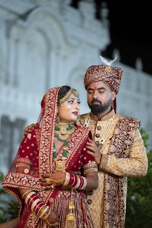 A couple dressed in traditional Indian wedding attire stands in an ornate setting. The woman is wearing a richly decorated red lehenga embellished with gold embroidery, along with intricate jewelry including a necklace, earrings, and bangles. The man is wearing a coordinated sherwani, featuring detailed embroidery and a turban. They appear poised and formal.