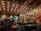 A cozy restaurant interior with low lighting and unique ceiling decorations featuring small bulbs and spring-like structures. The walls are adorned with various framed artworks and posters, creating a vibrant and eclectic atmosphere. There are several people seated at tables, enjoying their meals and drinks. The seating consists of colorful cushions and dark tables, contributing to the intimate and artistic ambiance.