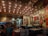 A cozy restaurant interior with low lighting and unique ceiling decorations featuring small bulbs and spring-like structures. The walls are adorned with various framed artworks and posters, creating a vibrant and eclectic atmosphere. There are several people seated at tables, enjoying their meals and drinks. The seating consists of colorful cushions and dark tables, contributing to the intimate and artistic ambiance.