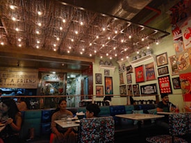 A cozy restaurant interior with low lighting and unique ceiling decorations featuring small bulbs and spring-like structures. The walls are adorned with various framed artworks and posters, creating a vibrant and eclectic atmosphere. There are several people seated at tables, enjoying their meals and drinks. The seating consists of colorful cushions and dark tables, contributing to the intimate and artistic ambiance.
