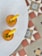 Two bright orange-colored drinks served in glasses are garnished with a lemon wedge and a sprig of mint, placed on a speckled white round table. The background reveals a patterned floor with geometric designs in colors of red, beige, blue, and white.