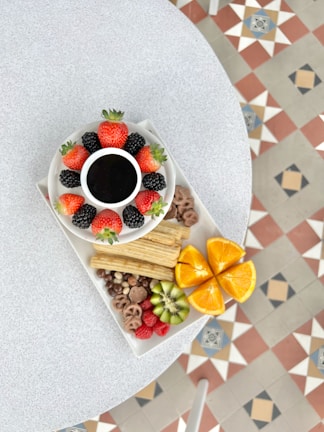 A round white table features a platter with various fruits and snacks. In the center is a small bowl of dark chocolate sauce surrounded by strawberries and blackberries. The platter also includes churros, halved oranges, sliced kiwi, raspberries, and mixed nuts with chocolate-covered pretzels, presenting a vibrant assortment of colors and textures. The geometric patterned floor tiles add a colorful backdrop.