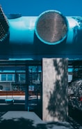 An outdoor area featuring a playground with a bright blue tube structure overhead, large grates, and a shadowed spiral staircase. The ground is shaded with various patterns, and a single swing is visible. The overall structure appears industrial with a modern design.