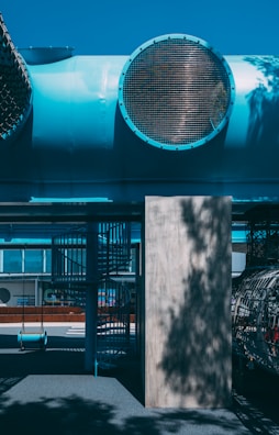 An outdoor area featuring a playground with a bright blue tube structure overhead, large grates, and a shadowed spiral staircase. The ground is shaded with various patterns, and a single swing is visible. The overall structure appears industrial with a modern design.