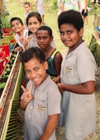 A group of children, some wearing school uniforms, are gathered around a small garden with potted plants. They are smiling and making peace signs, suggesting a cheerful and lively atmosphere. The background shows a grassy area with more children visible, possibly at a school or community event.