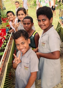 A group of children, some wearing school uniforms, are gathered around a small garden with potted plants. They are smiling and making peace signs, suggesting a cheerful and lively atmosphere. The background shows a grassy area with more children visible, possibly at a school or community event.