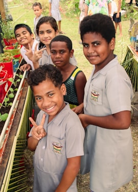 A group of children, some wearing school uniforms, are gathered around a small garden with potted plants. They are smiling and making peace signs, suggesting a cheerful and lively atmosphere. The background shows a grassy area with more children visible, possibly at a school or community event.