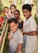 A group of children, some wearing school uniforms, are gathered around a small garden with potted plants. They are smiling and making peace signs, suggesting a cheerful and lively atmosphere. The background shows a grassy area with more children visible, possibly at a school or community event.