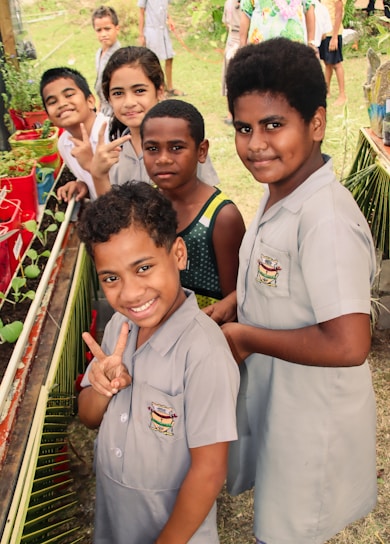 A group of children, some wearing school uniforms, are gathered around a small garden with potted plants. They are smiling and making peace signs, suggesting a cheerful and lively atmosphere. The background shows a grassy area with more children visible, possibly at a school or community event.