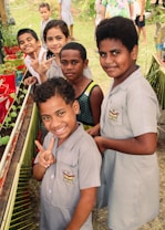A group of children, some wearing school uniforms, are gathered around a small garden with potted plants. They are smiling and making peace signs, suggesting a cheerful and lively atmosphere. The background shows a grassy area with more children visible, possibly at a school or community event.