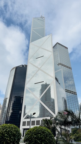 A modern skyscraper with a steel and glass facade features a distinctive cross-bracing design. The structure is surrounded by other high-rise buildings and lush greenery at the base. The sky is partly cloudy, creating a dramatic backdrop.