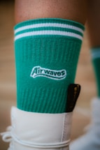 A close-up of a green sock with white stripes around the top, featuring the word 'Airwaves' in white text. The sock is worn with a white shoe.