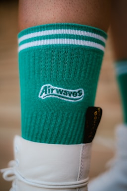 A close-up of a green sock with white stripes around the top, featuring the word 'Airwaves' in white text. The sock is worn with a white shoe.