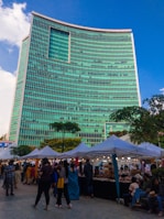 A bustling street market set up along the side of a towering modern building with a striking turquoise-colored facade. The market stalls, covered with white canopies, are lined up in neat rows and attract a diverse crowd of people browsing various goods. The lively scene is illuminated by small hanging lights under the canopies, creating a festive atmosphere. Trees and greenery add a touch of nature to the urban setting.