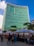 A bustling street market set up along the side of a towering modern building with a striking turquoise-colored facade. The market stalls, covered with white canopies, are lined up in neat rows and attract a diverse crowd of people browsing various goods. The lively scene is illuminated by small hanging lights under the canopies, creating a festive atmosphere. Trees and greenery add a touch of nature to the urban setting.