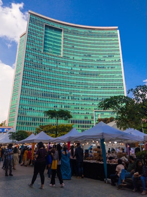 A bustling street market set up along the side of a towering modern building with a striking turquoise-colored facade. The market stalls, covered with white canopies, are lined up in neat rows and attract a diverse crowd of people browsing various goods. The lively scene is illuminated by small hanging lights under the canopies, creating a festive atmosphere. Trees and greenery add a touch of nature to the urban setting.