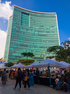 A bustling street market set up along the side of a towering modern building with a striking turquoise-colored facade. The market stalls, covered with white canopies, are lined up in neat rows and attract a diverse crowd of people browsing various goods. The lively scene is illuminated by small hanging lights under the canopies, creating a festive atmosphere. Trees and greenery add a touch of nature to the urban setting.